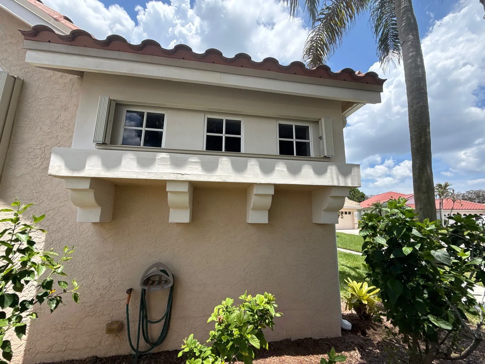Exterior Painting in Port Saint Lucie, Florida. Single-storey stucco exterior in light beige with a white corbelled window ledge, white-trimmed windows and barrel-tile roof edge, surrounded by shrubs and a palm.