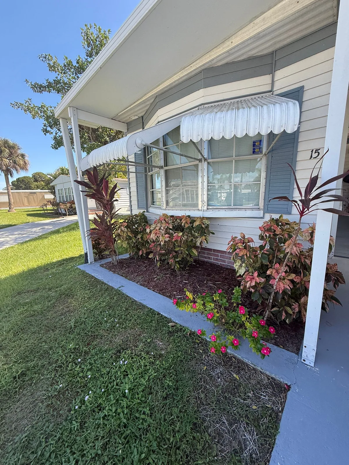 Exterior Painting in Port Saint Lucie, Florida. Manufactured home exterior with pale blue siding, white porch posts, striped window awning and planted bed of flowering shrubs.