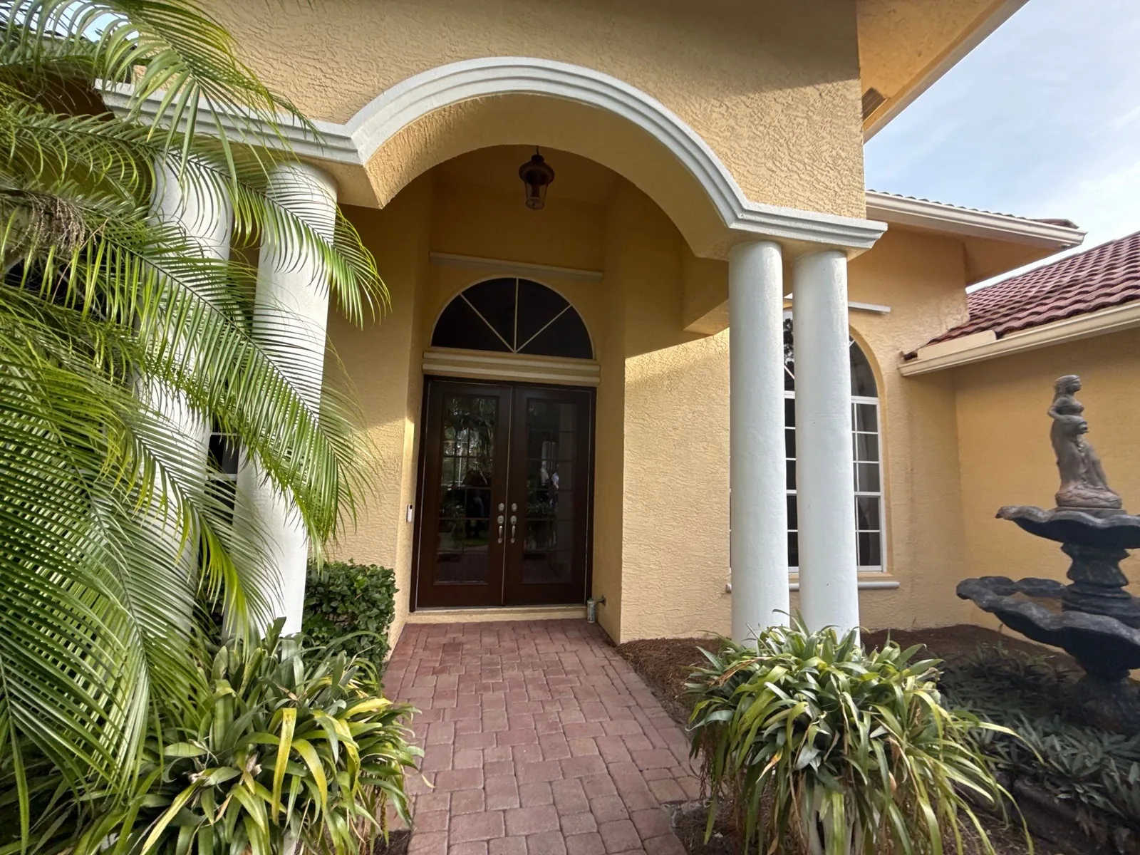 Exterior Painting in Port Saint Lucie, Florida. Arched entry portico on a yellow stucco home with white columns and trim, dark wood double doors and brick paver walkway.