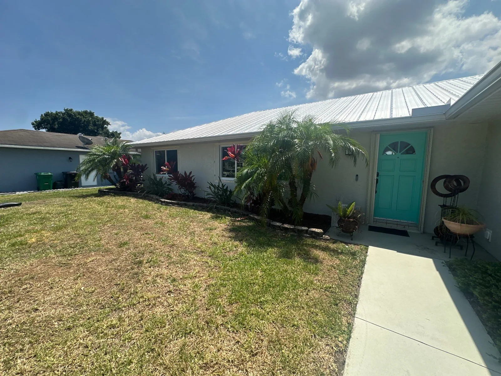 Exterior Painting in Port Saint Lucie, Florida. Front and side elevation of a single-storey house with light stucco walls, white trim and a teal front door.