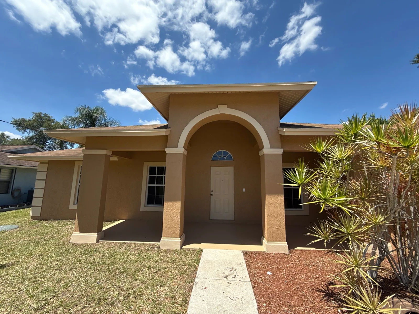 Exterior Painting in Port Saint Lucie, Florida. Single-storey tan stucco house front elevation with arched entry portico, white door and columns, under blue sky.