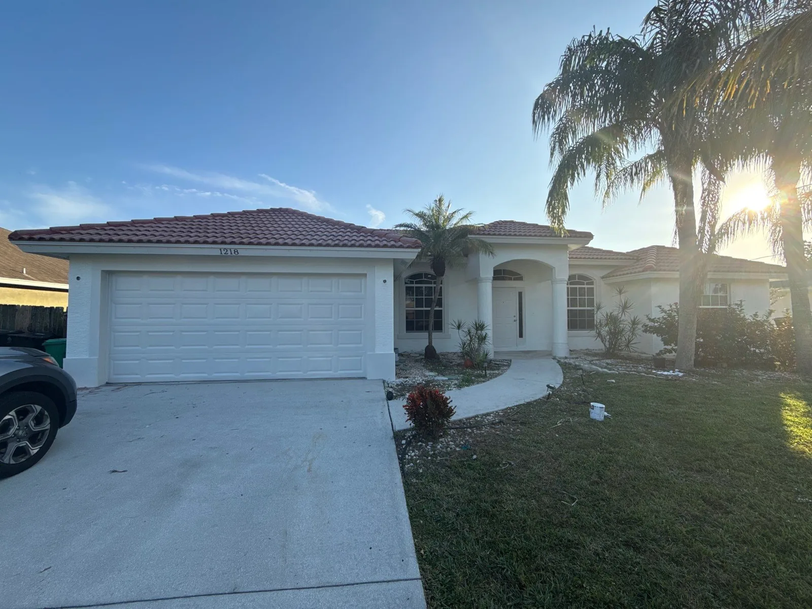 Exterior Painting in Port Saint Lucie, Florida. Front elevation of a single-storey white stucco house with barrel tile roof, white two-car garage door and palm trees.