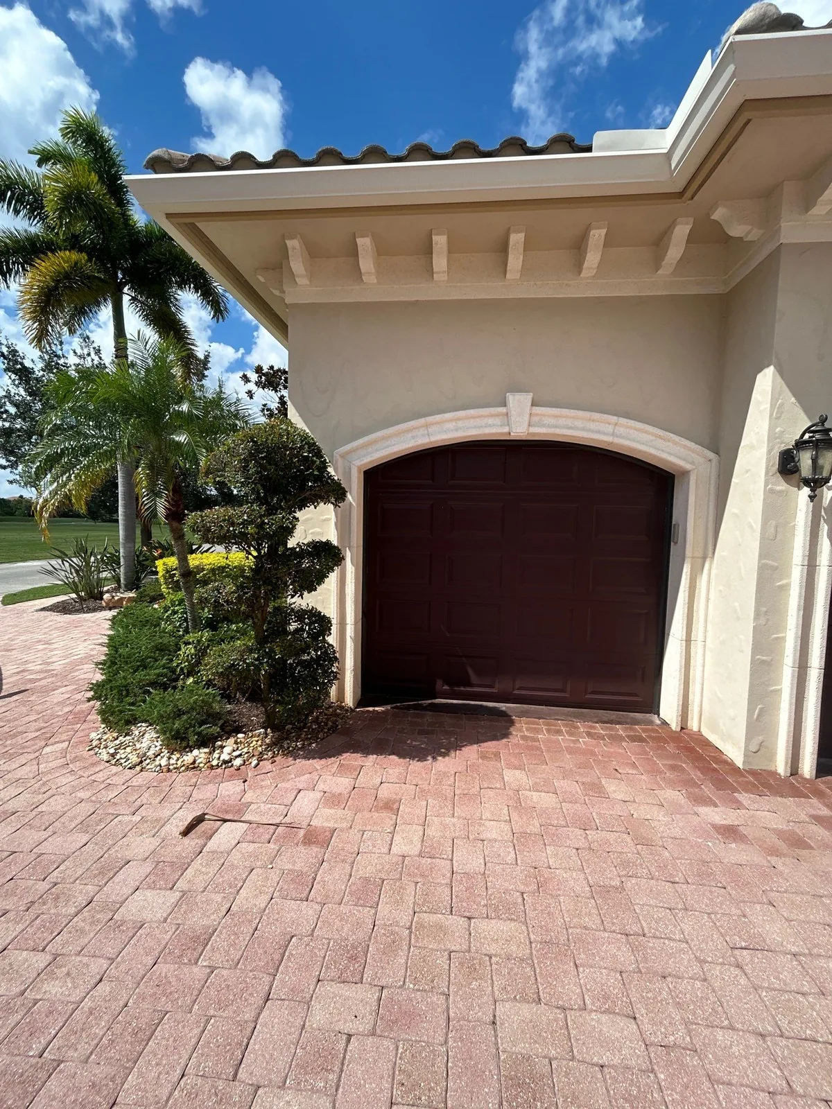Exterior Painting in Port Saint Lucie, Florida. Arched garage bay on a stucco home with a dark panelled door, trimmed shrubs, palms and a paver driveway.