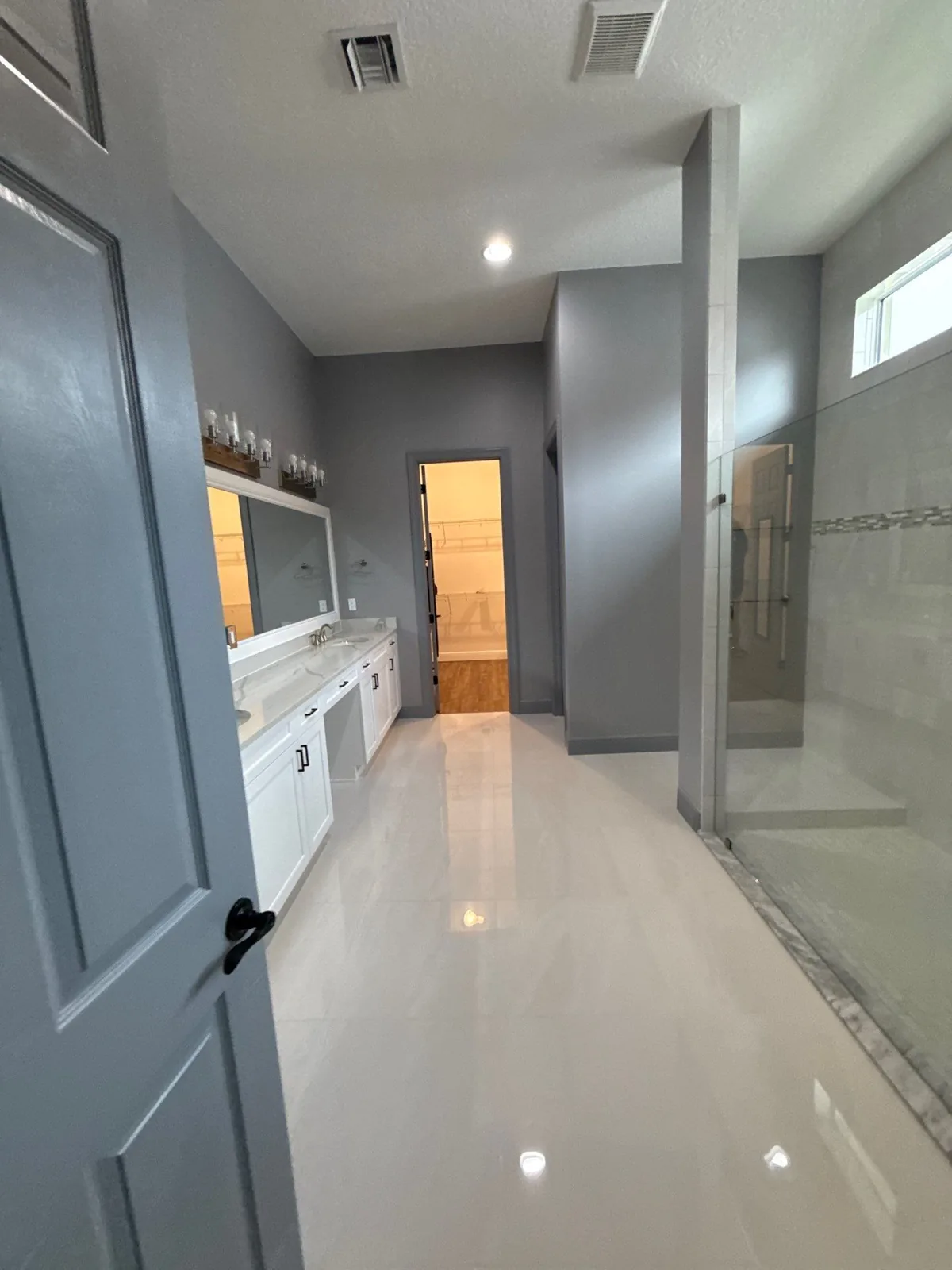 Interior Painting in Port Saint Lucie, Florida. Long master bathroom with grey walls, white double vanity, glass shower enclosure and glossy tile floor.