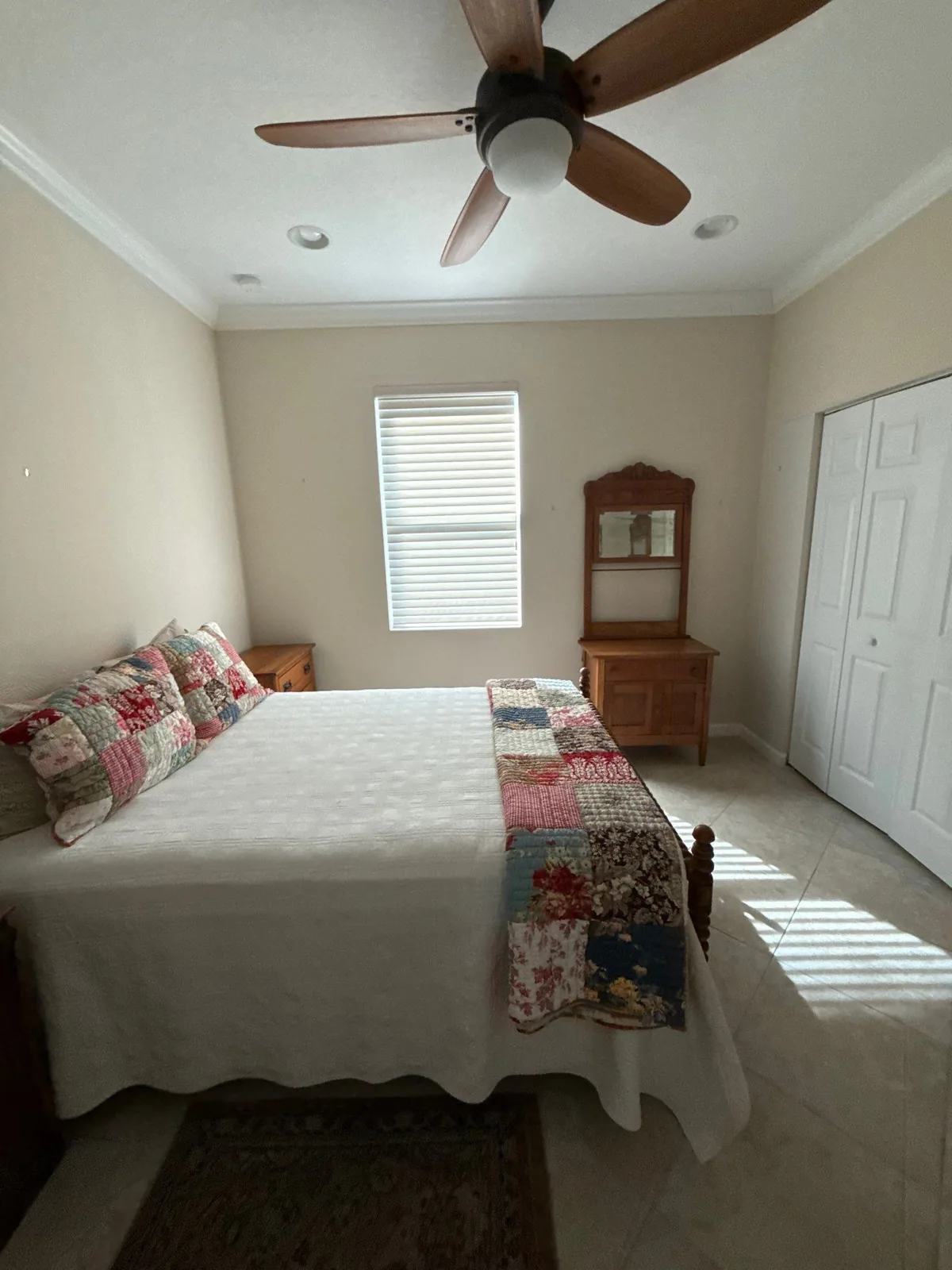 Interior Painting in Port Saint Lucie, Florida. Bedroom with cream painted walls, white crown moulding and door trim, ceiling fan and a made bed by a shuttered window.