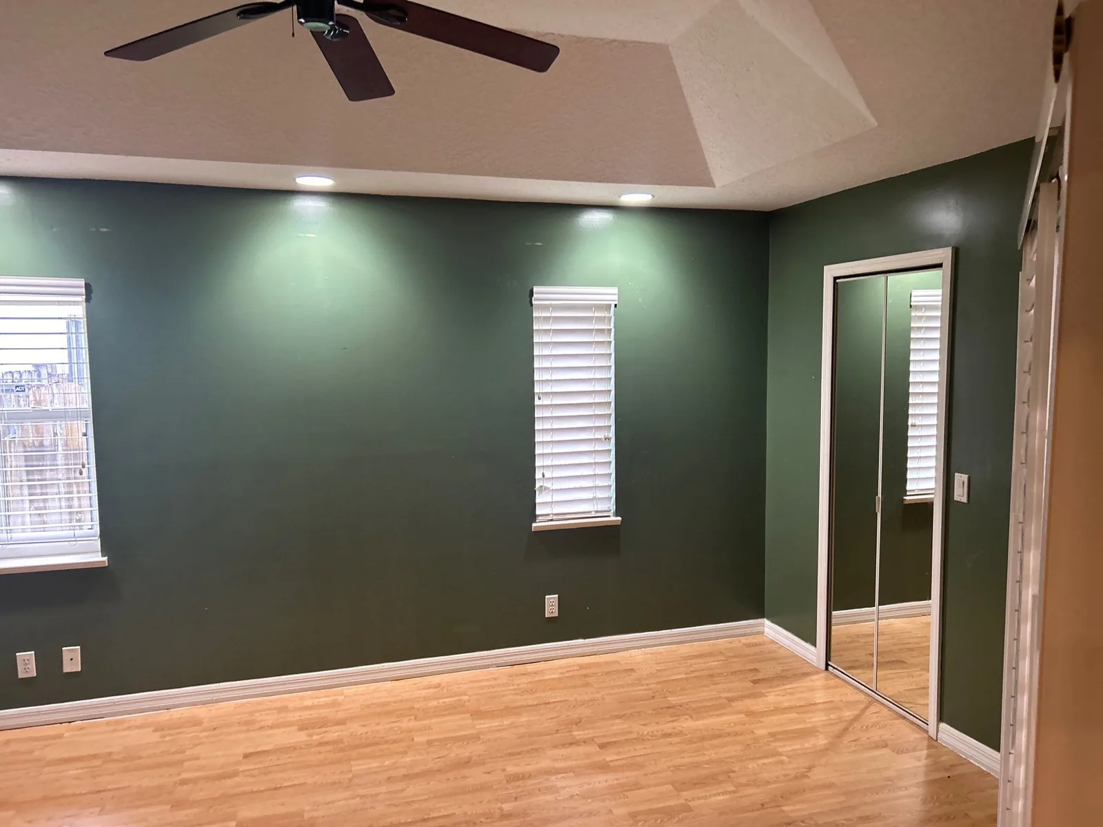 Interior Painting in Port Saint Lucie, Florida. Empty bedroom with dark green painted walls, white trim and baseboards, mirrored closet doors, ceiling fan and laminate flooring.