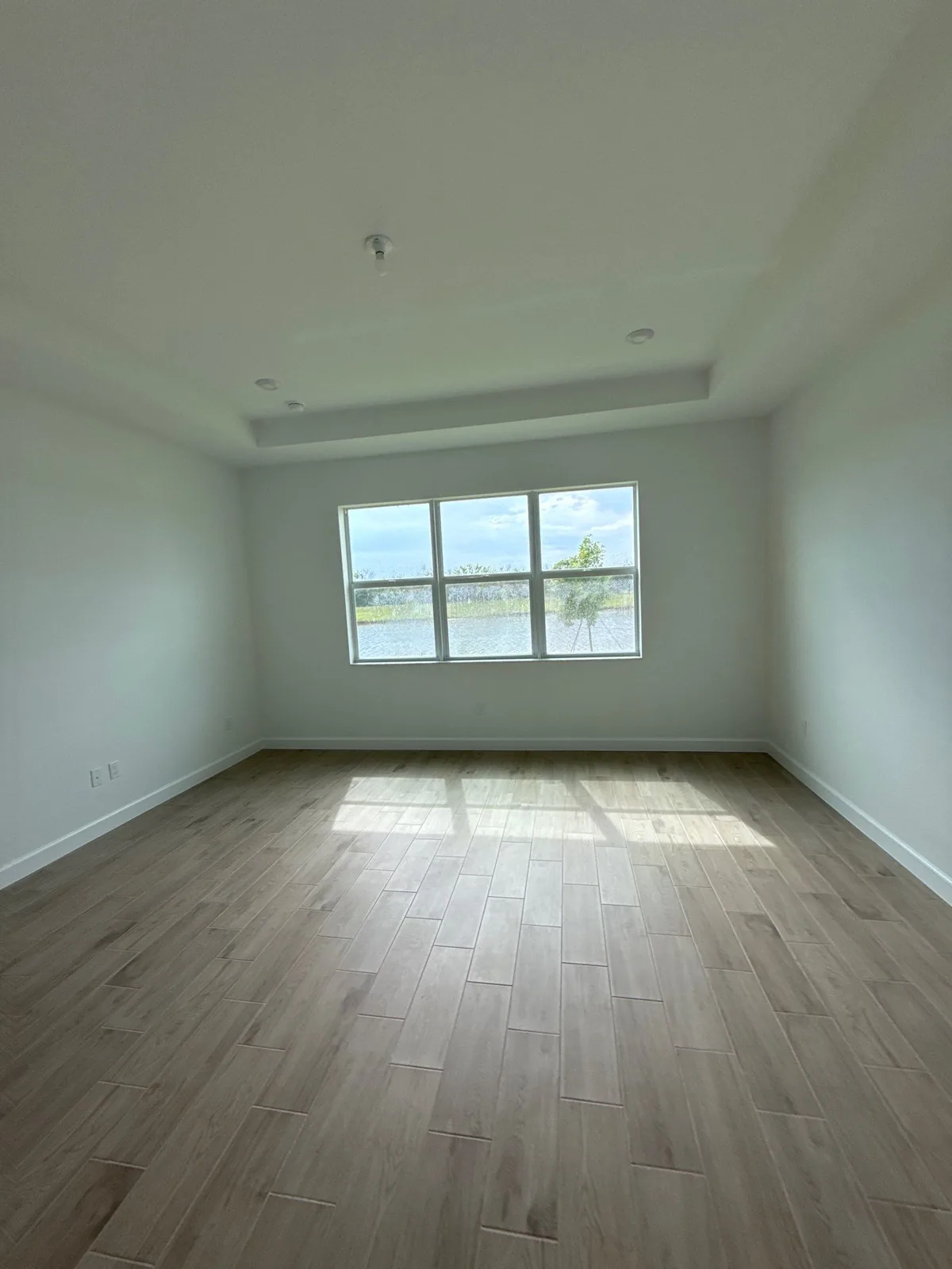 Interior Painting in Port Saint Lucie, Florida. Empty bedroom with white walls and tray ceiling, white baseboards, wood-look plank flooring and a large window.