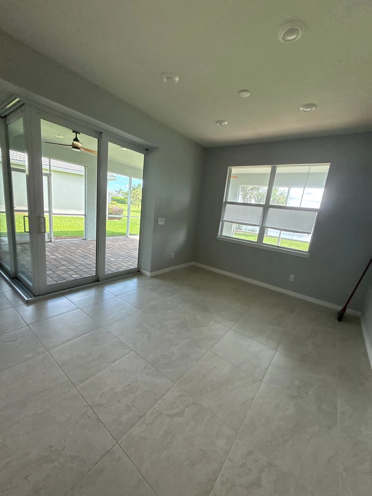 Interior Painting in Port Saint Lucie, Florida. Empty room with grey walls and white ceiling and baseboards, sliding glass doors to a covered patio and two shaded windows, large tile floor.