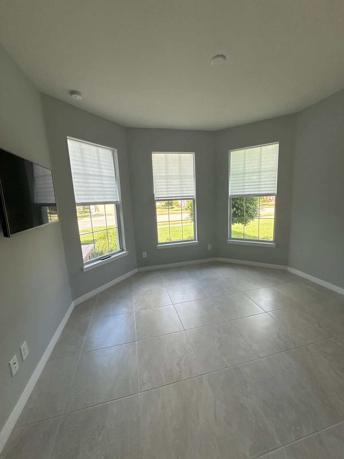 Interior Painting in Port Saint Lucie, Florida. Empty room with light grey walls and white trim, three shaded windows in a bay, tile floor and a wall-mounted television.