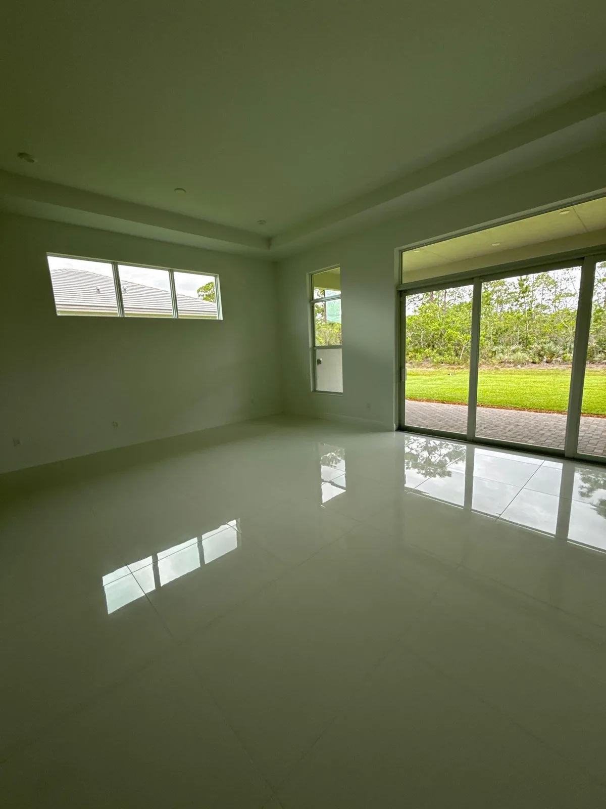 Interior Painting in Port Saint Lucie, Florida. Empty room with pale green-grey painted walls, white tray ceiling and trim, polished tile floor and a sliding glass door onto a lawn.