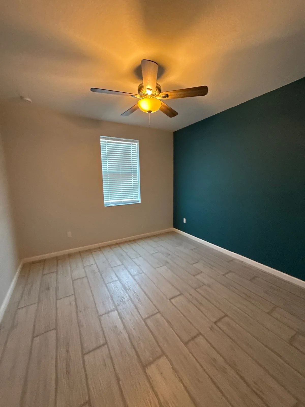 Interior Painting in Port Saint Lucie, Florida. Empty bedroom with tan walls and a dark teal accent wall, lit ceiling fan and a window with blinds.