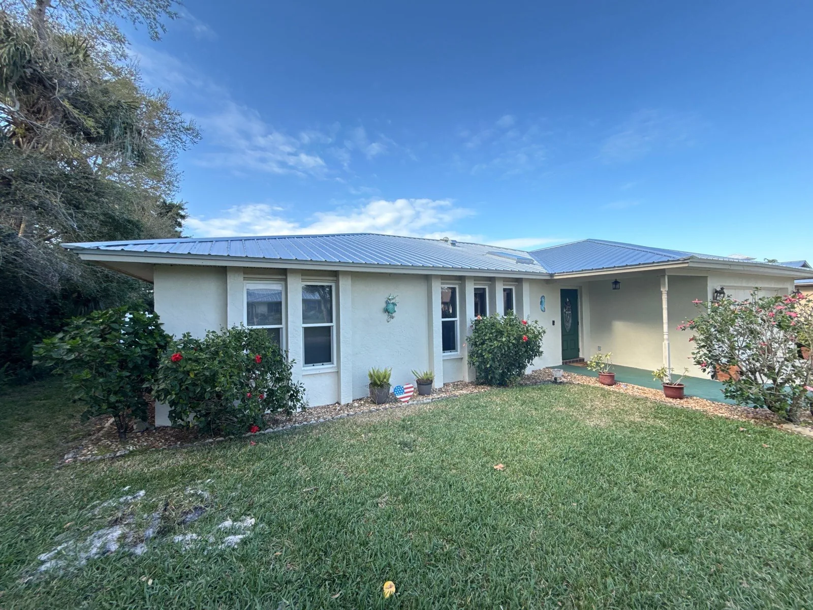 Exterior Painting in Port Salerno, Florida. Single-storey beige stucco house with white trim, blue metal roof, lawn and flowering shrubs under a blue sky.