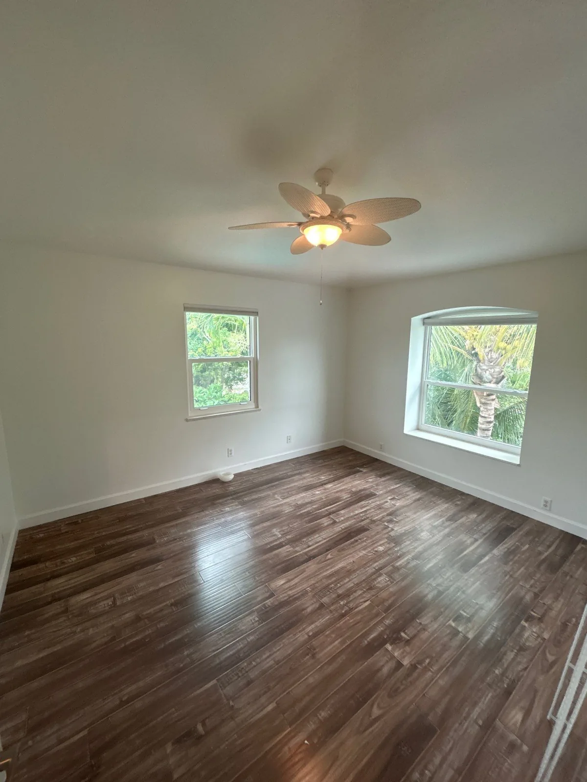 Interior Painting in Port Salerno, Florida. Empty bedroom with white painted walls and ceiling, two windows, a ceiling fan and dark wood-look plank flooring.
