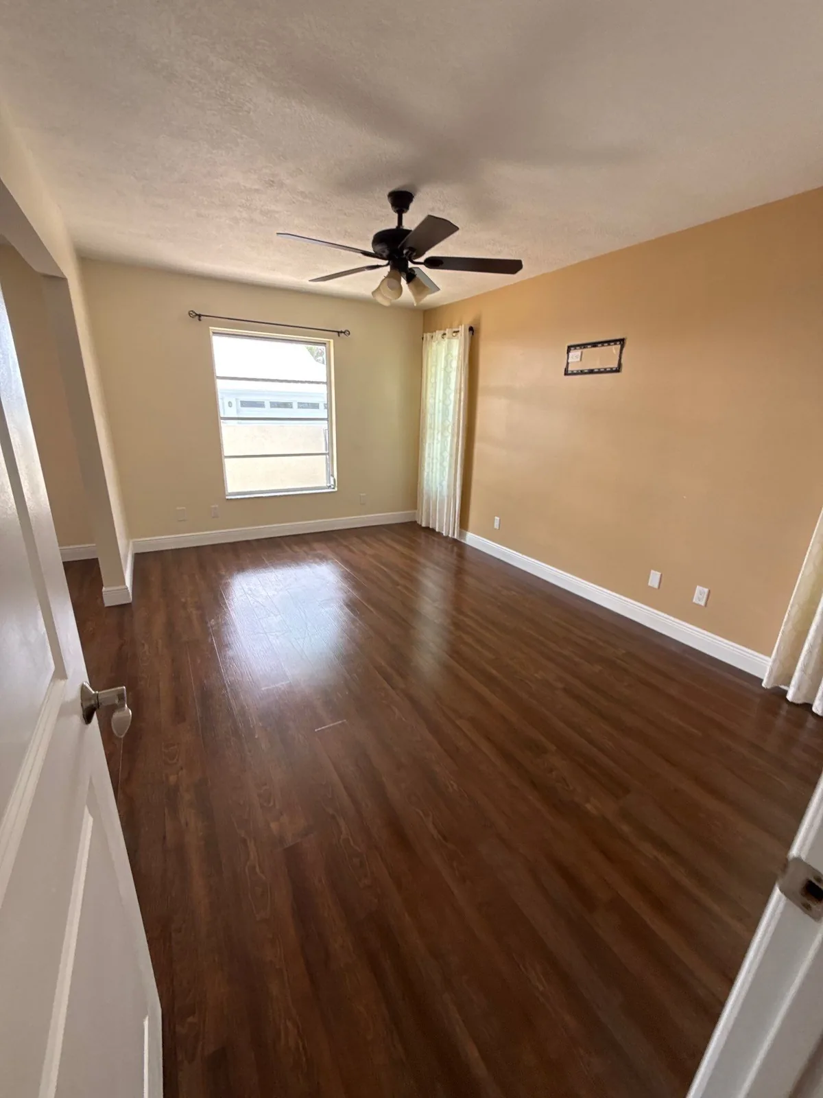 Interior Painting in Port Salerno, Florida. Empty bedroom with tan walls, white ceiling and trim, ceiling fan, curtained window and dark wood-look flooring.