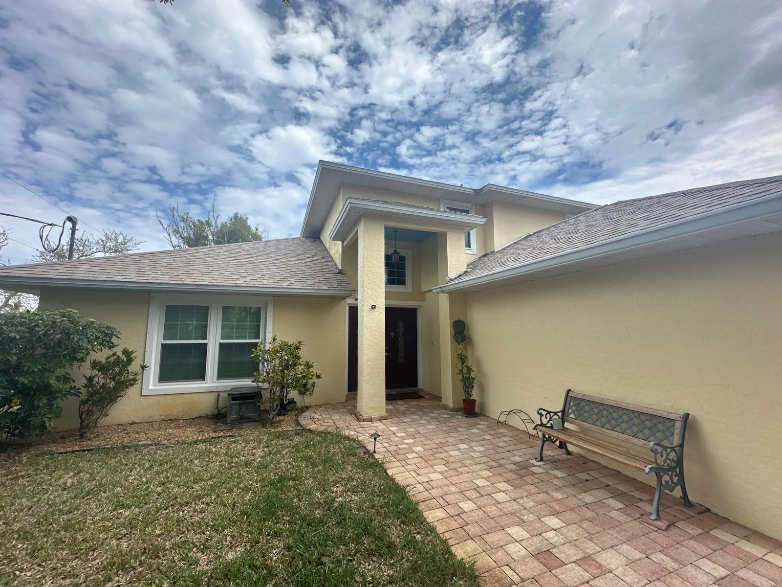 Exterior Painting in Roseland, Florida. Yellow stucco home entry with white window trim, shingle roof, paver walkway and a metal bench under a blue sky.