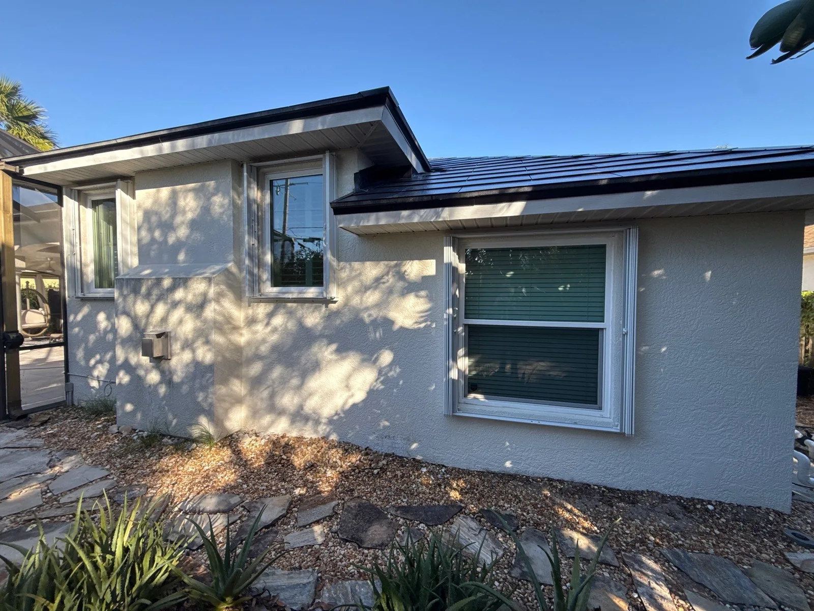 Exterior Painting in Satellite Beach, Florida. Single-storey stucco house exterior in light neutral with white window trim and dark metal roof.