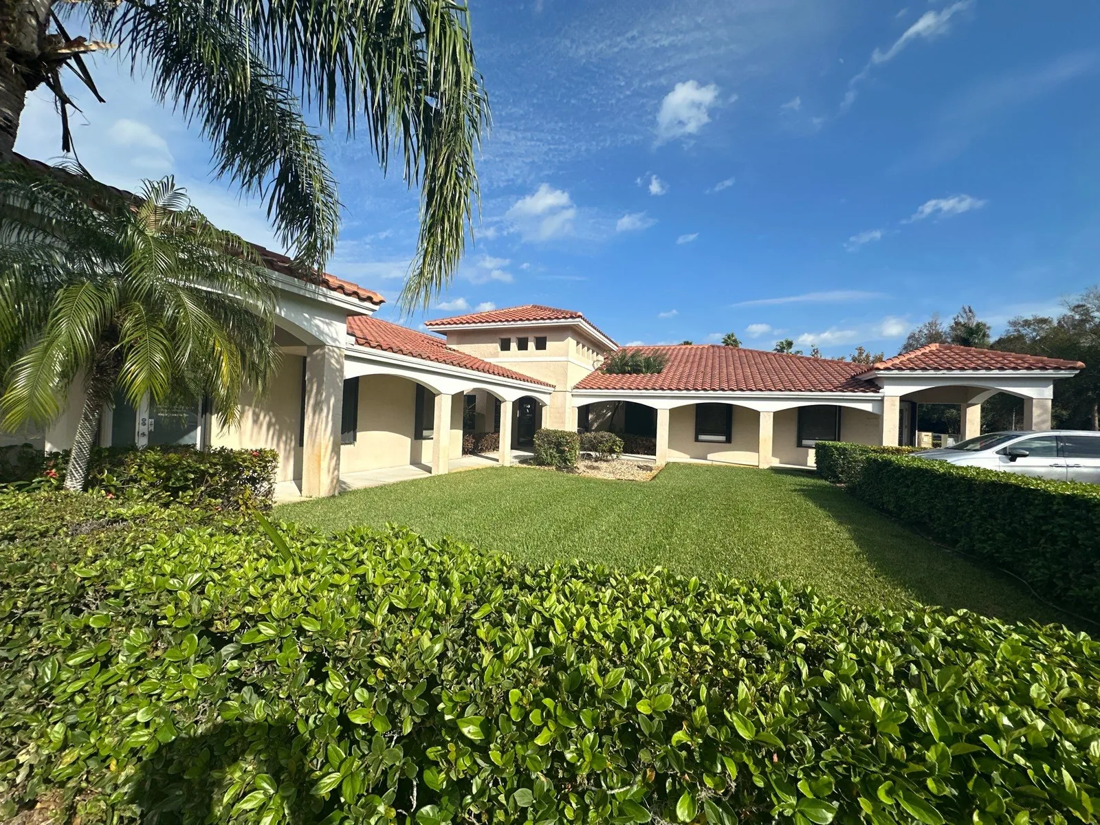 Exterior Painting in Stuart, Florida. Single-storey home with red barrel tile roof and cream stucco walls, arched columned porch, lawn and clipped hedges under a blue sky.