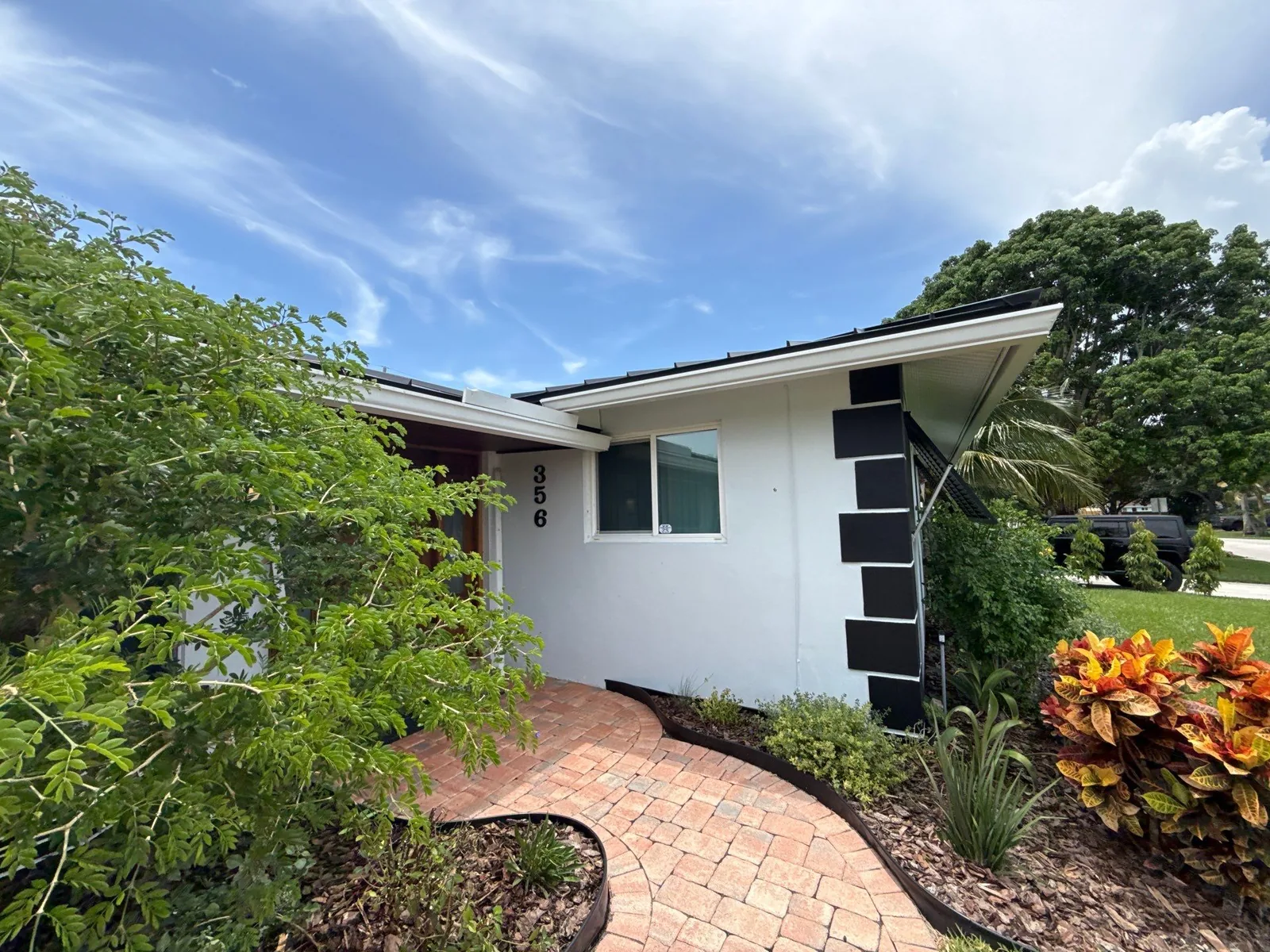Exterior Painting in Tequesta, Florida. Single-storey house with freshly painted white stucco, black accent panels, brick paver walkway and landscaping under a blue sky.