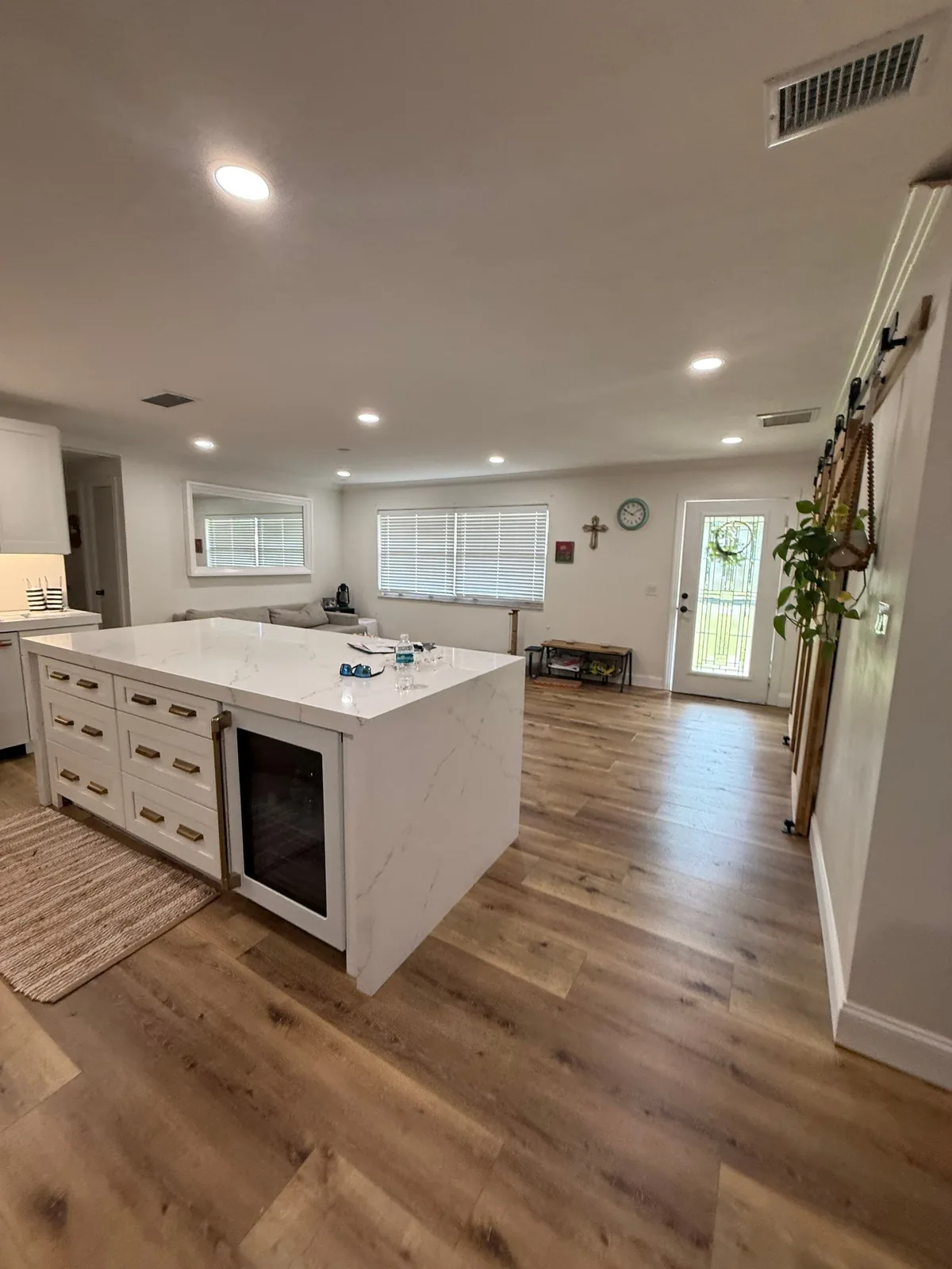 Interior Painting in Tequesta, Florida. Open-plan kitchen and living area with white walls and ceiling, white island with stone countertop and wine fridge, wood-look plank flooring.