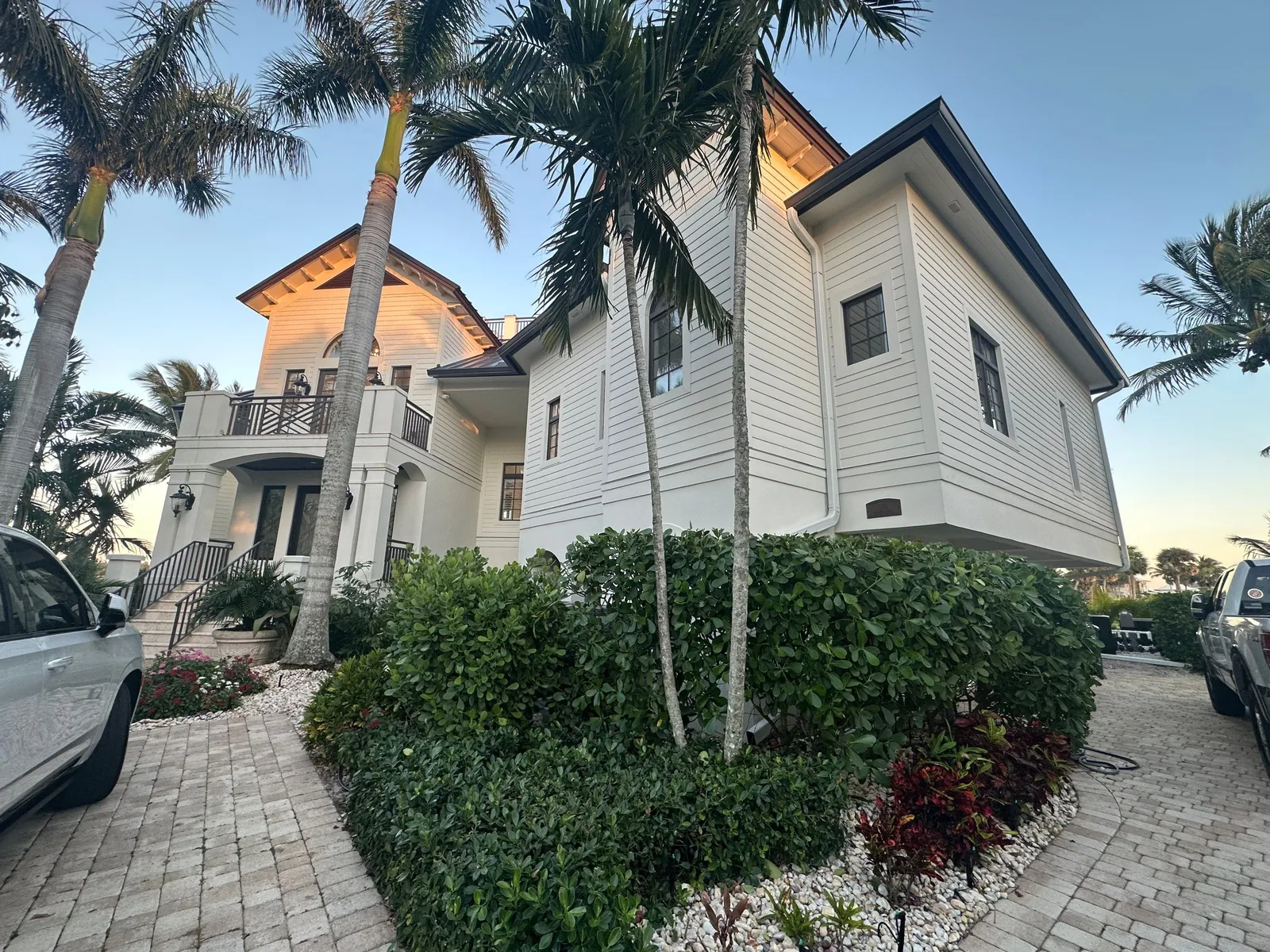 Exterior Painting in Vero Beach, Florida. Two-storey white lap-siding home with dark trim and metal roof accents, palm trees and hedged landscaping in front.