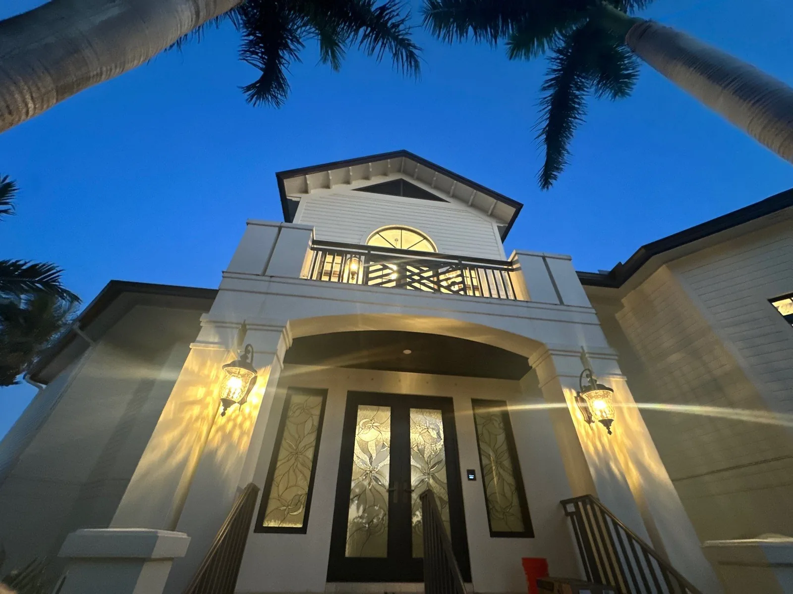 Exterior Painting in Vero Beach, Florida. Front entry of a white two-storey home at dusk with glass double doors, lit wall sconces, arched portico and upper balcony.