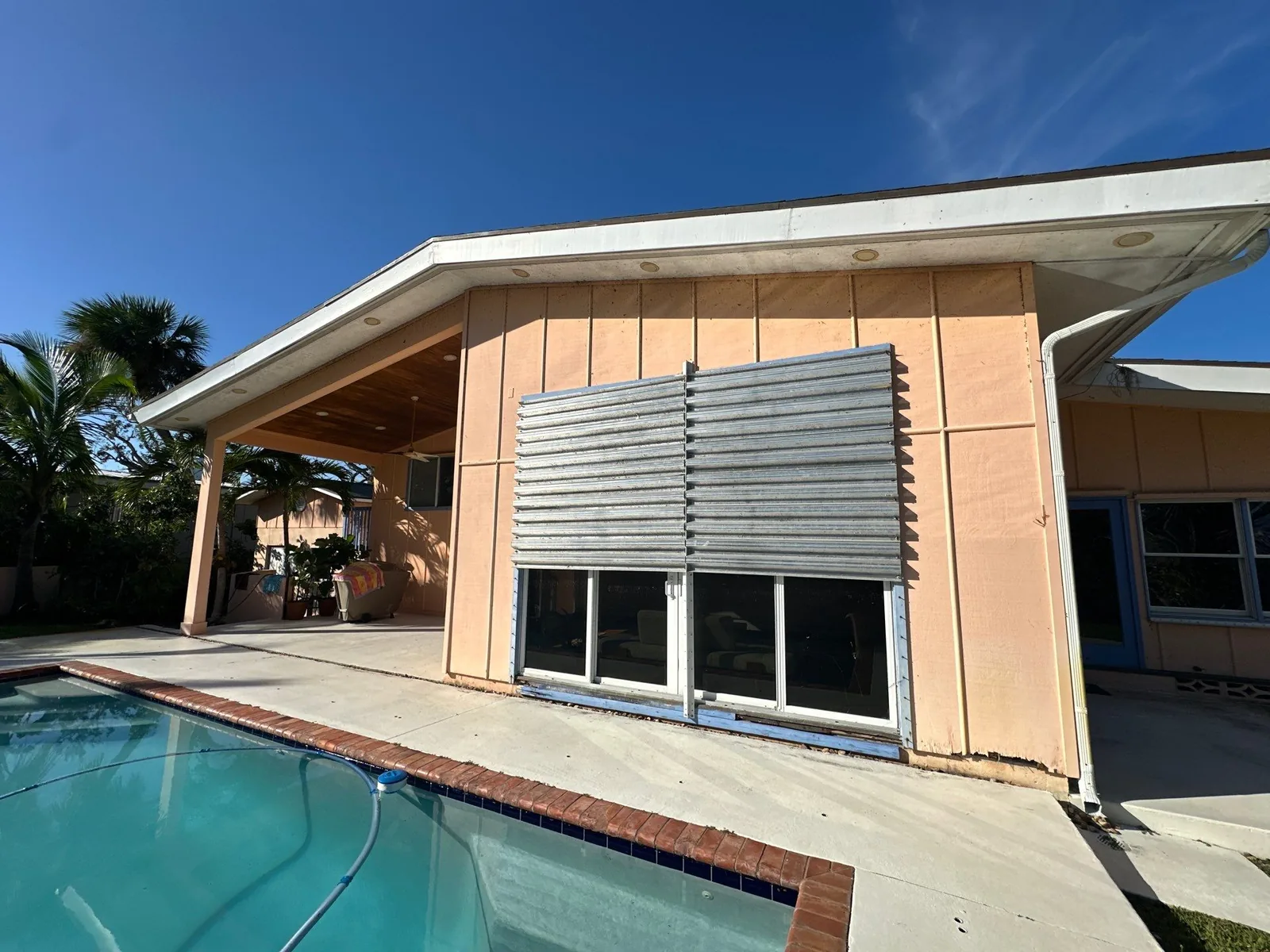 Exterior Painting in Vero Beach, Florida. Rear elevation of a home with peach painted walls and white fascia, sliding glass doors and metal shutters beside a swimming pool.