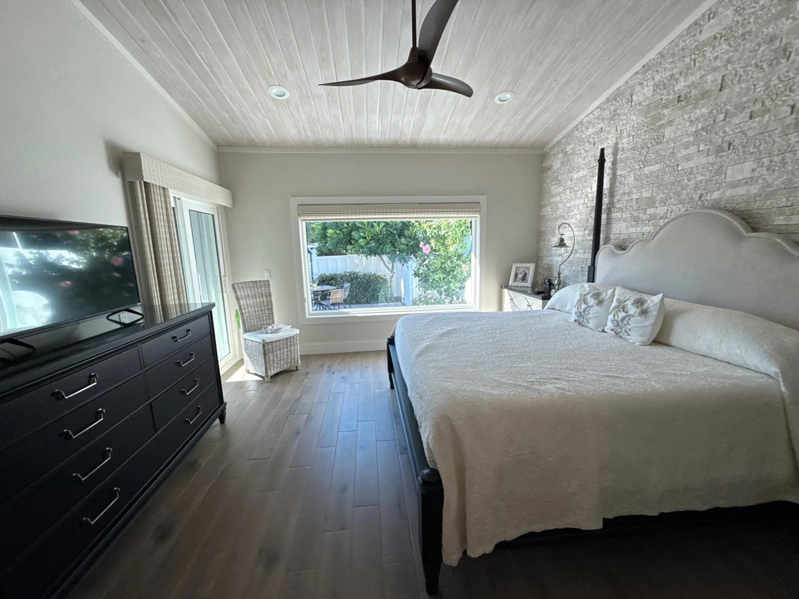 Interior Painting in Vero Beach, Florida. Bedroom with a white painted tongue-and-groove vaulted ceiling, pale walls, a stone veneer accent wall, a large window and dark wood flooring.
