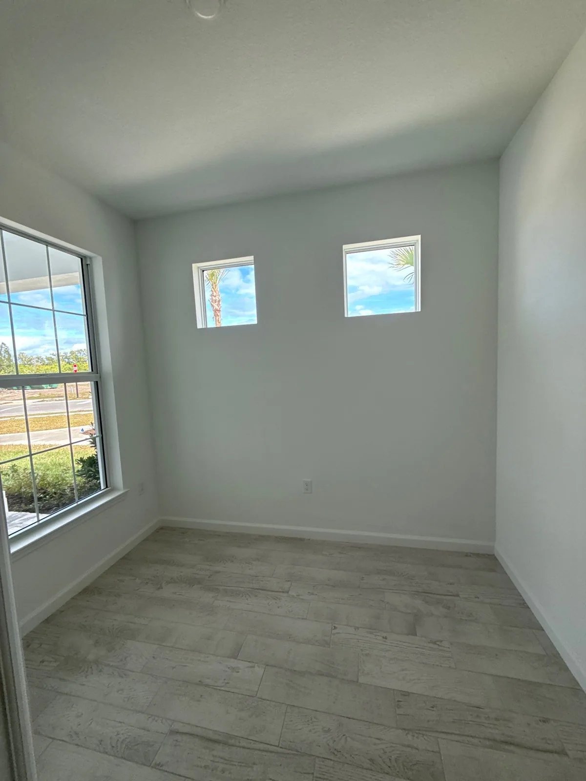 Interior Painting in Wabasso, Florida. Empty room with white walls, two small high windows and a large corner window, grey plank flooring.