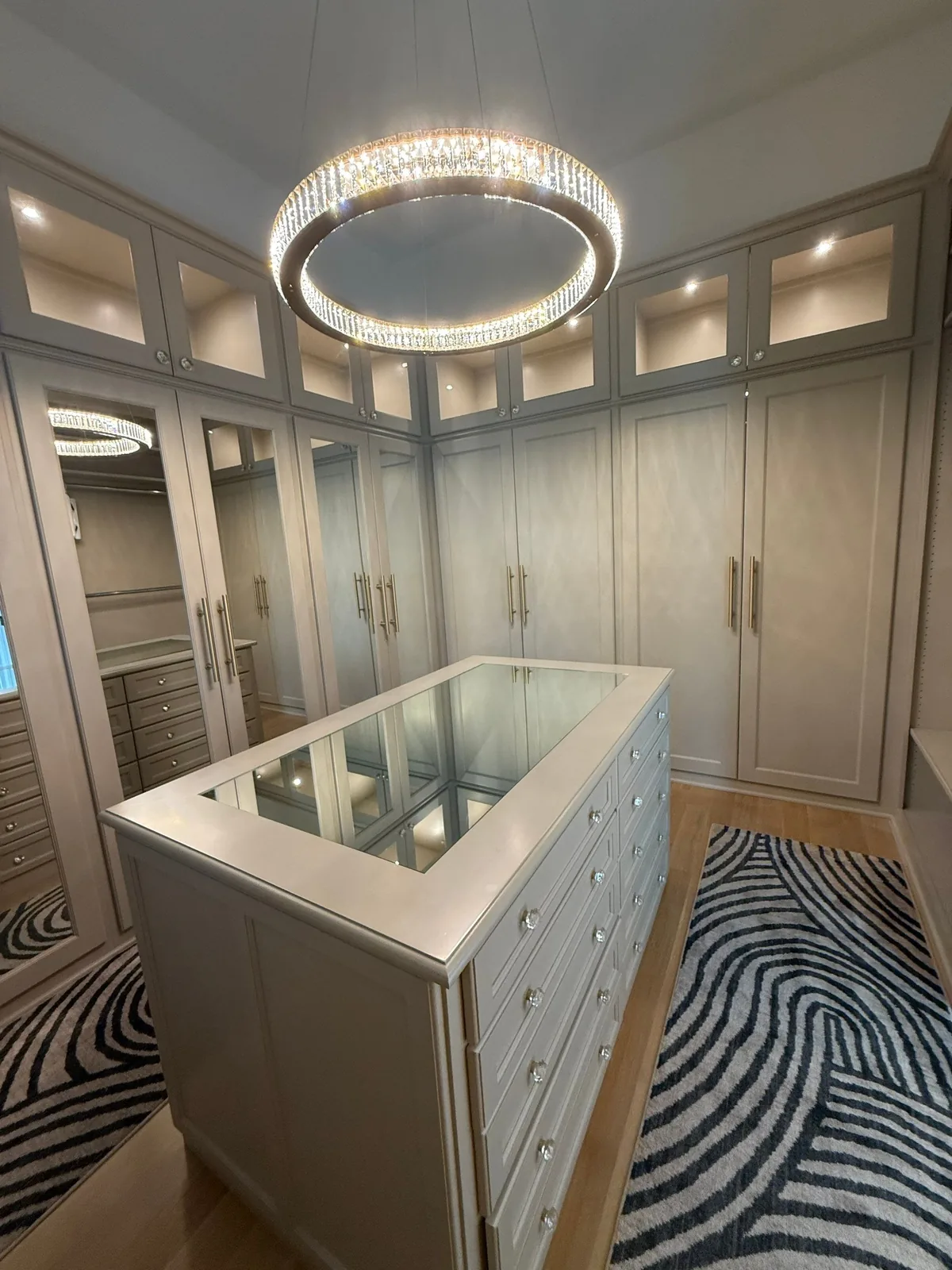 Cabinet Refinishing in Wellington, Florida. Walk-in closet with light neutral built-in cabinetry on three walls, a glass-topped centre island of drawers and a round crystal ceiling fixture.