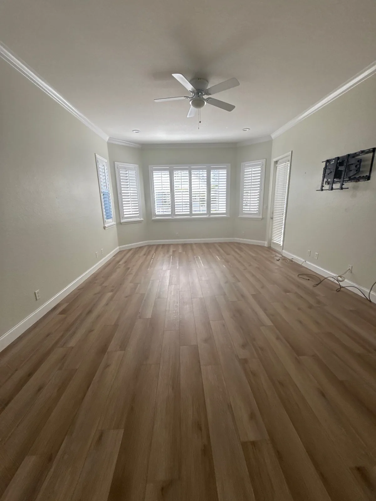 Interior Painting in Wellington, Florida. Empty living room with cream walls, white crown moulding and baseboards, plantation shutters on three windows, wood-look plank flooring and a ceiling fan.