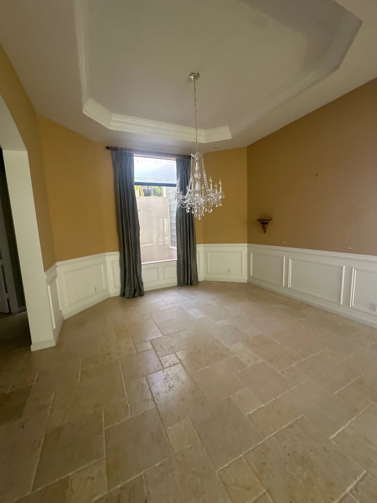 Interior Painting in Whisper Walk, Florida. Empty dining room with tray ceiling, warm tan painted walls above white wainscot panelling, crystal chandelier and curtained window.