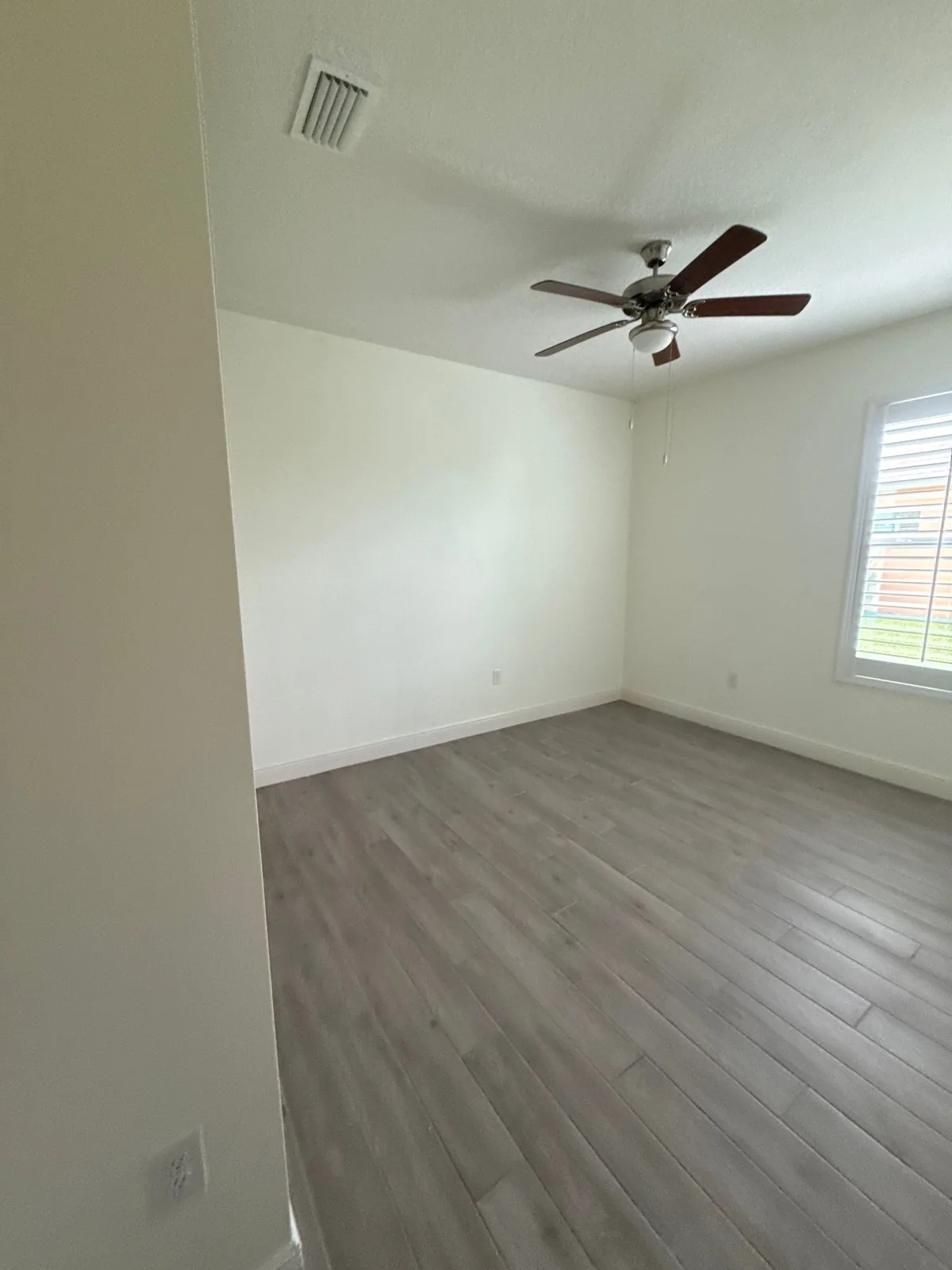 Interior Painting in Winter Beach, Florida. Empty bedroom with freshly painted white walls and ceiling, ceiling fan, shuttered window and grey wood-look plank flooring.